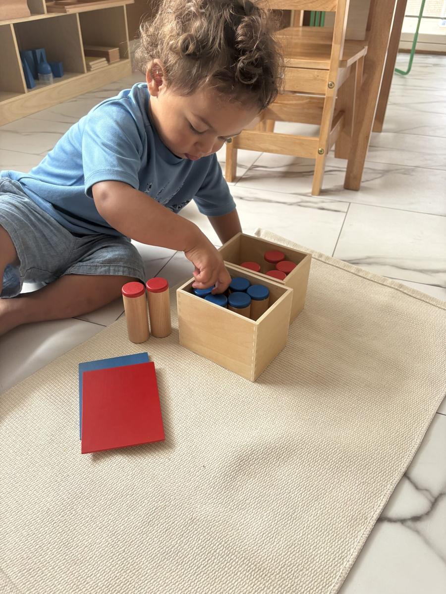 A toddler kneeling on a mat, reaching into a wooden box of red and blue discs to sort them beside matching red and blue cards