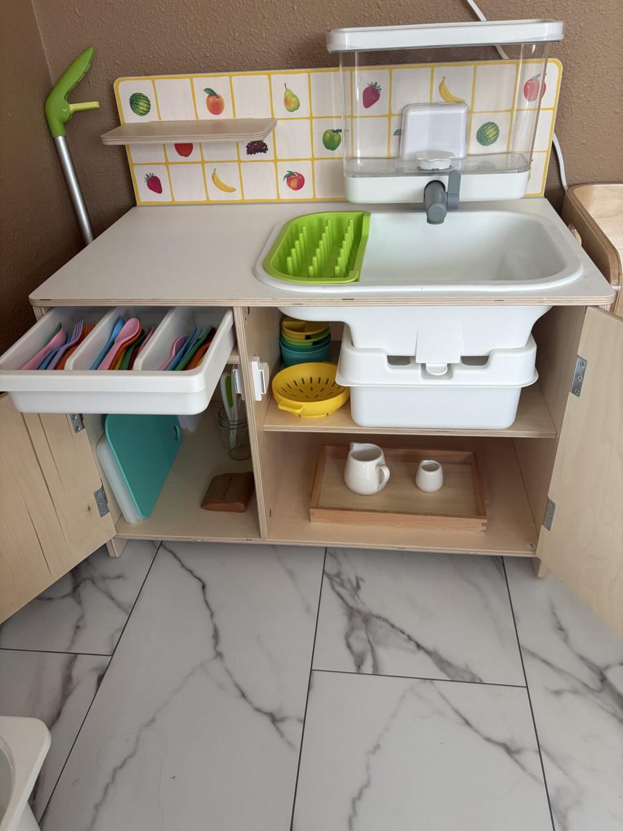 A child-height washing station with a basin, drying rack, and open shelves holding real cups, bowls, and cutting boards