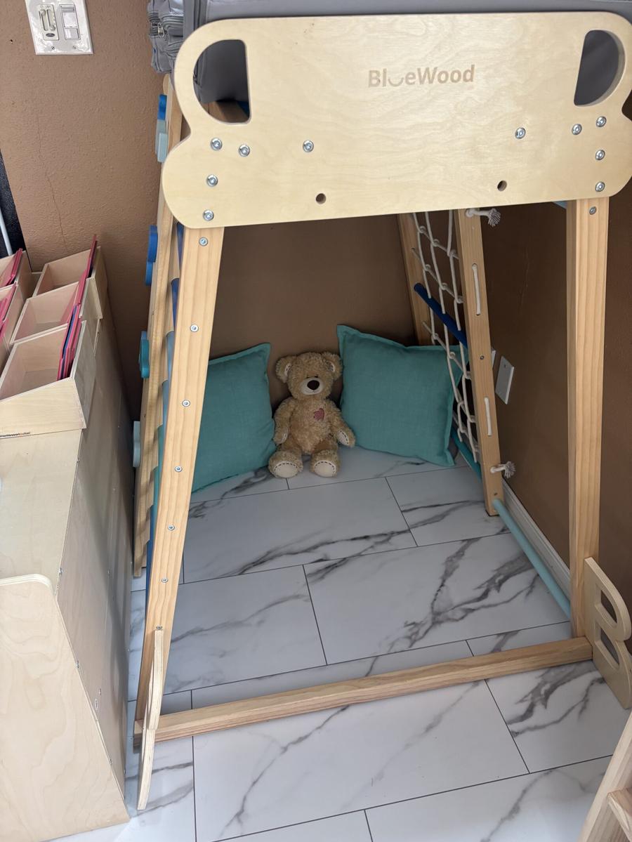 A wooden climbing frame turned into a reading nook with a cushioned mat, two green pillows, and a teddy bear