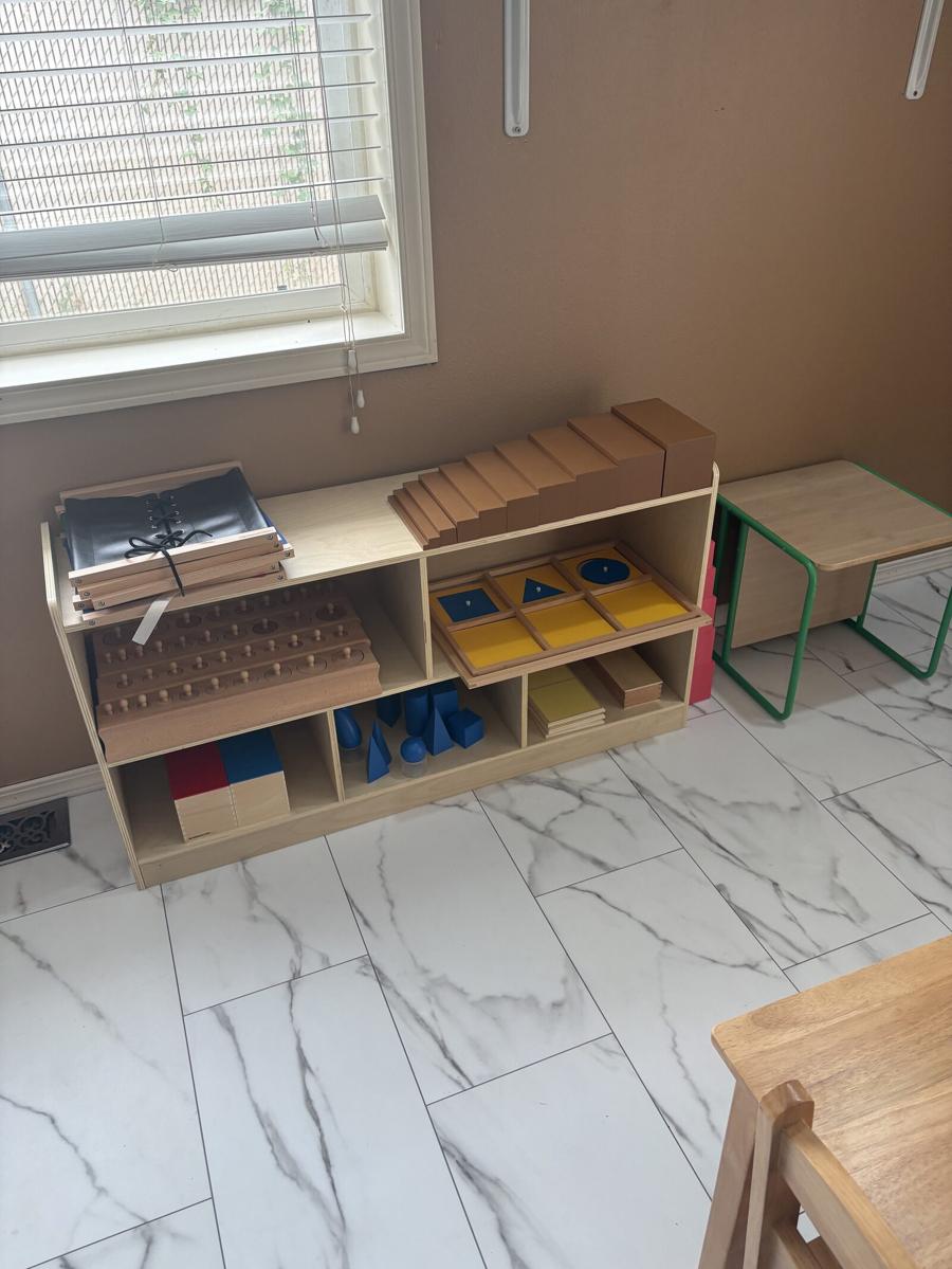 A sensorial shelf holding the brown stair, geometric cabinet drawers, and wooden geometric solids beside a window