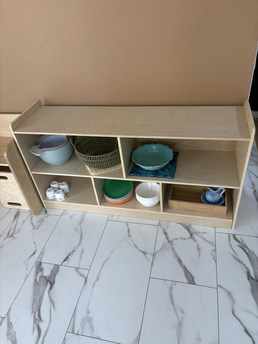 An open wooden shelf holding stacked ceramic bowls, plates, a woven basket, small pitchers, and teacups within a child's reach